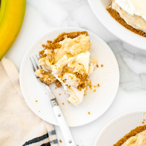 rustic slice of banana cream pie with graham cracker crust on a plate with a fork on a white marble counter.