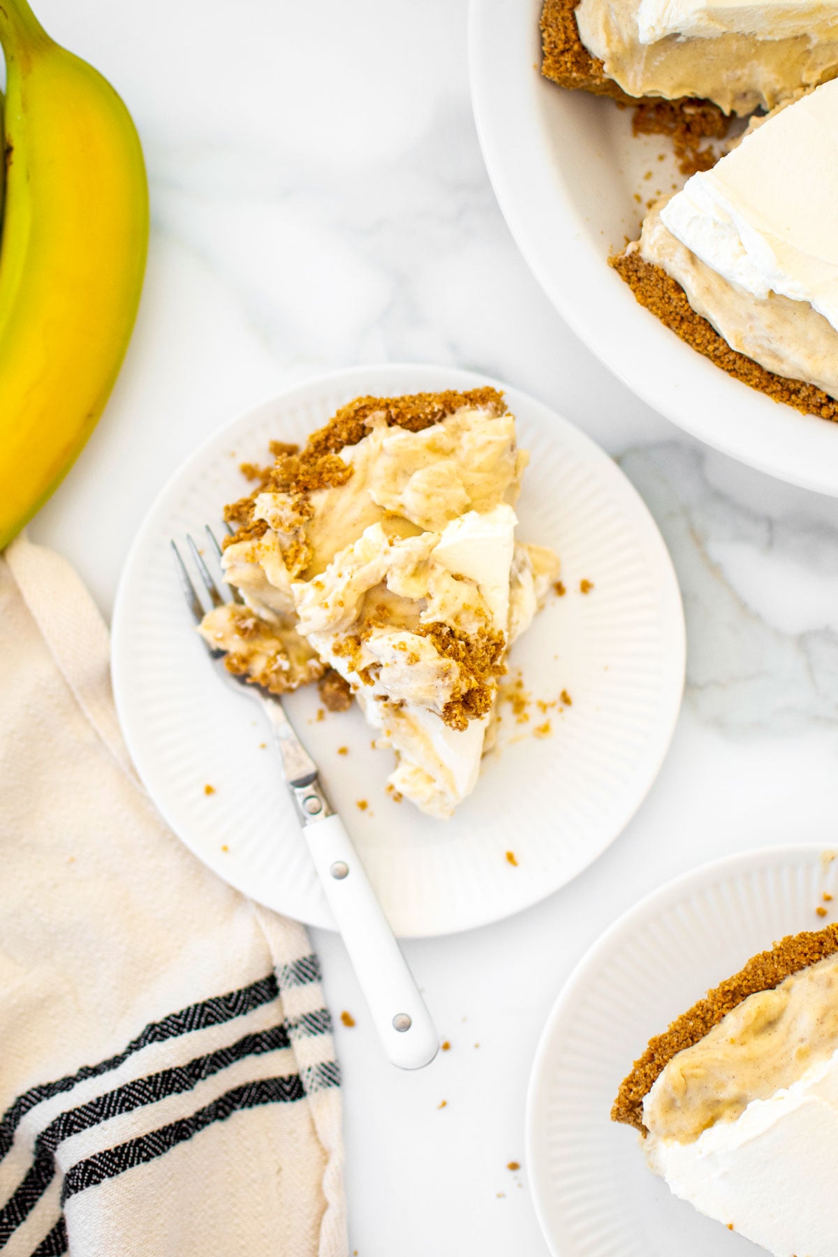 rustic slice of banana cream pie with graham cracker crust on a plate with a fork on a white marble counter.