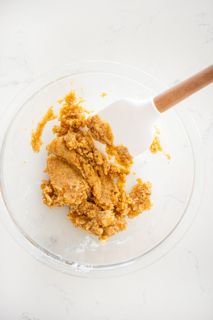 mixed crumble topping in a glass mixing bowl with a spatula on a marble counter.