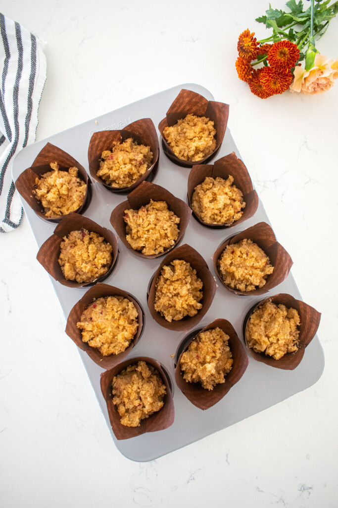 muffin pan with eggless banana raspberry muffin batter with crumble topping on top on a marble counter.