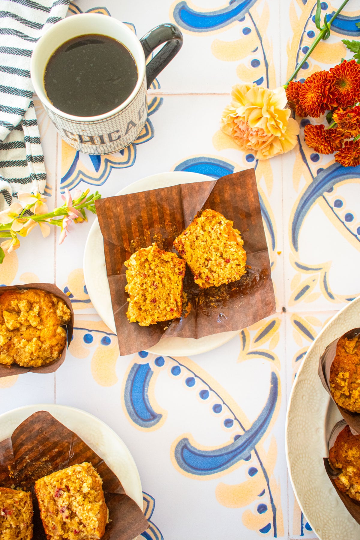 eggless banana raspberry muffin cut in half on a small plate on a tile table with a mug of coffee and flowers.