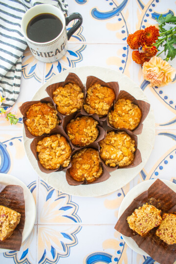 round plate of eggless banana raspberry muffins on a tile table with a mug of coffee and flowers.