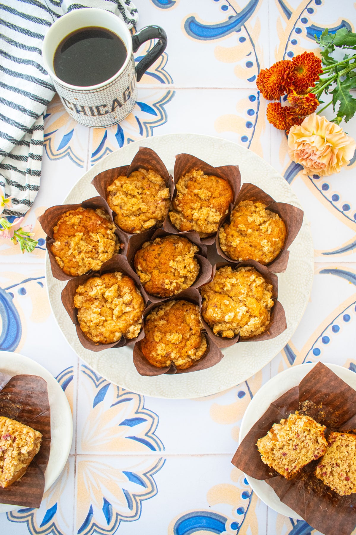 round plate of eggless banana raspberry muffins on a tile table with a mug of coffee and flowers.