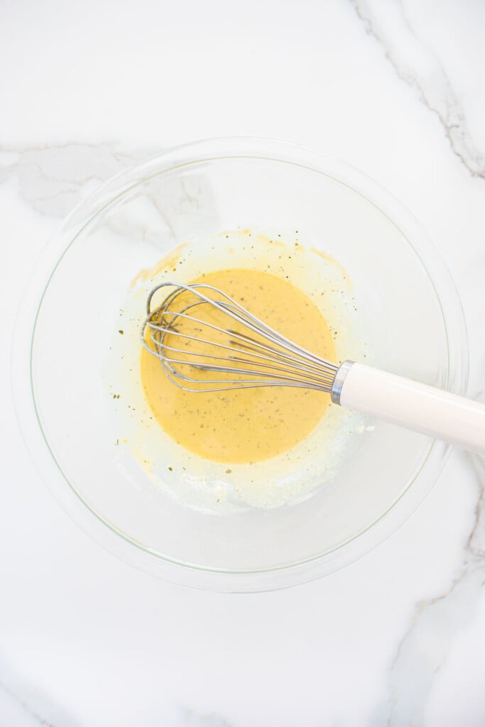 garlic powder, ranch seasoning and white cheddar powder in a glass bowl mixed with olive oil with a whisk on a white marble counter.