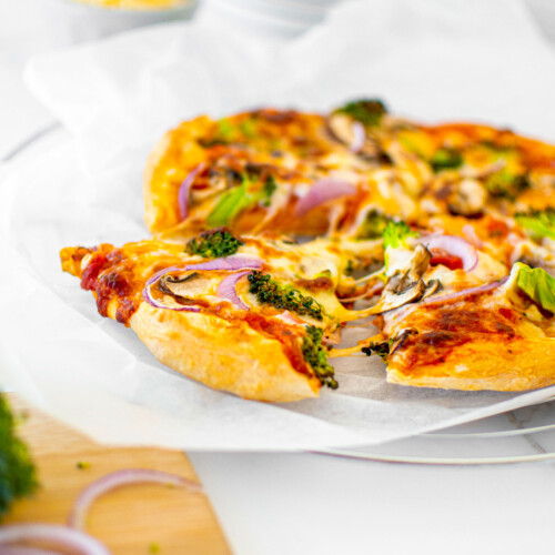 mushroom broccoli pizza slice being pulled out of a pie on parchment paper on a white marble countertop.