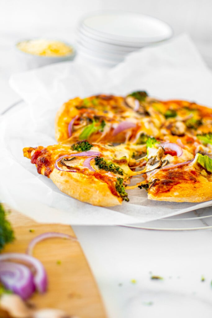 mushroom broccoli pizza slice being pulled out of a pie on parchment paper on a white marble countertop.