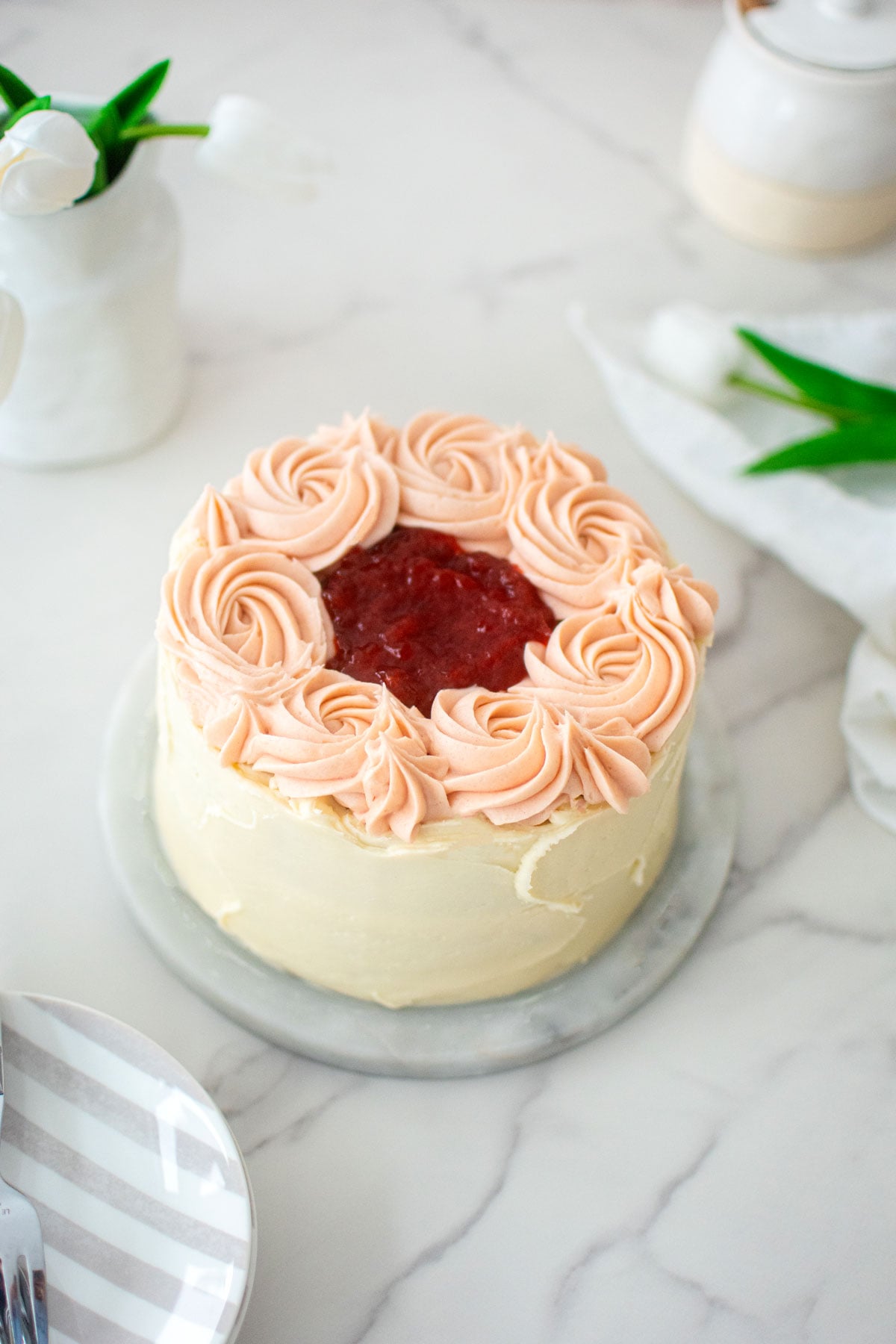 strawberry almond cake on a marble turntable with pink roses and strawberry jam in the center on a marble counter.