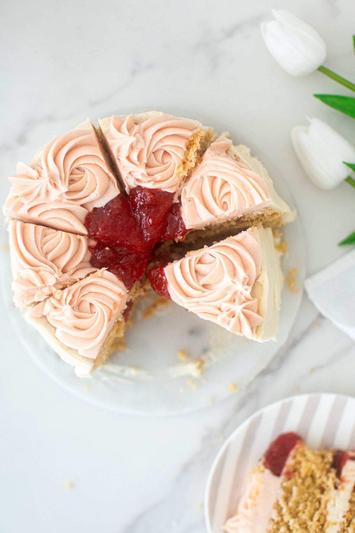 strawberry almond cake cut into slices on a marble turntable with pink roses and strawberry jam in the center on a marble counter.