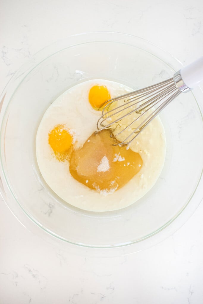 wet ingredients for almond cake in a glass mixing bowl on a marble counter with a whisk.