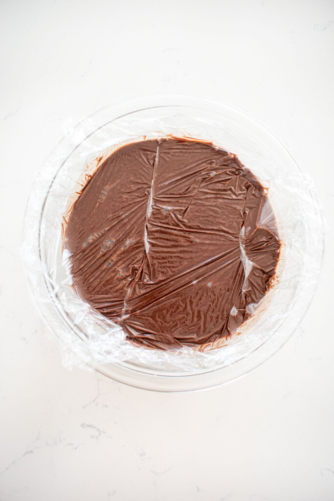 chocolate protein pudding in a glass mixing bowl with plastic wrap pressed into it.