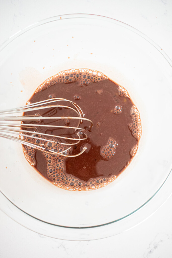 egg yolks mixed with chocolate pudding mixture in a glass mixing bowl with a whisk on a white marble counter.