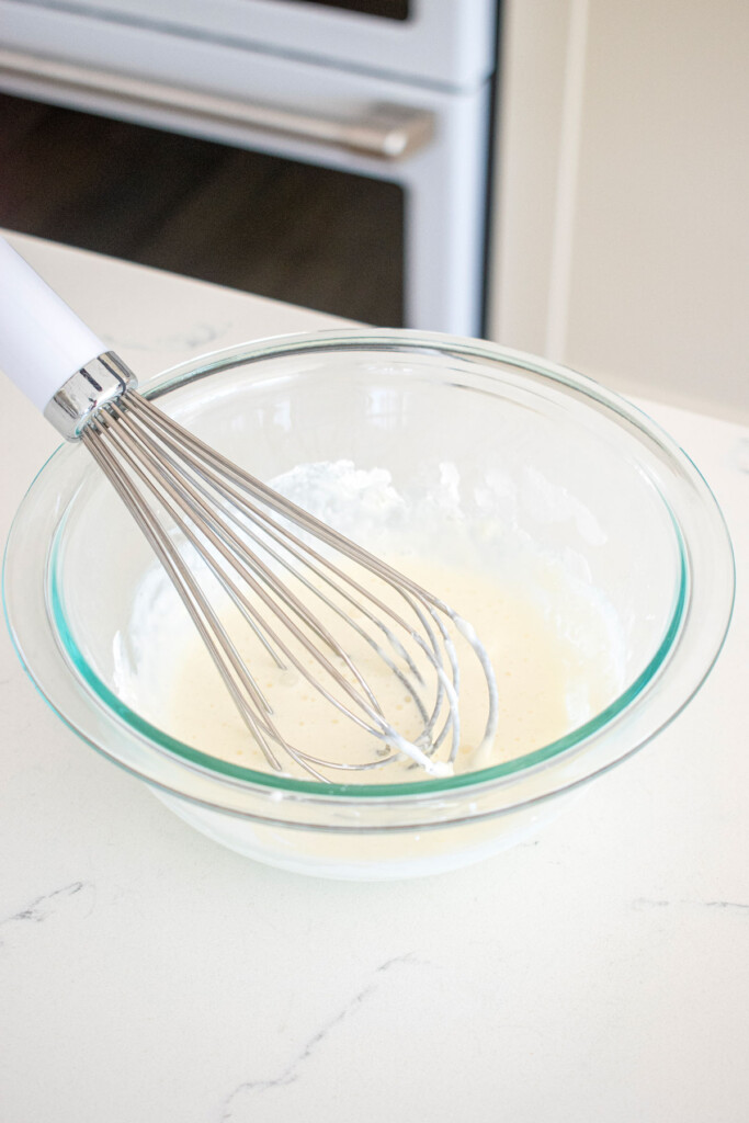 glass mixing bowl with cream and gelatin with a whisk on a marble counter.