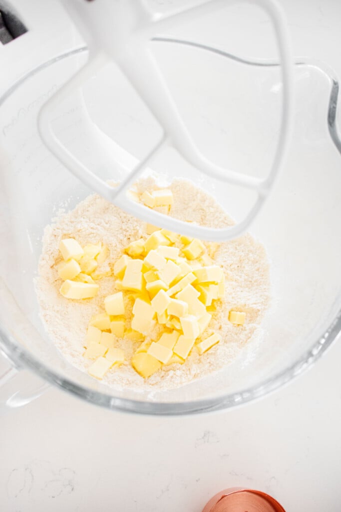 chopped butter added to a glass mixing bowl with flour on a marble counter.
