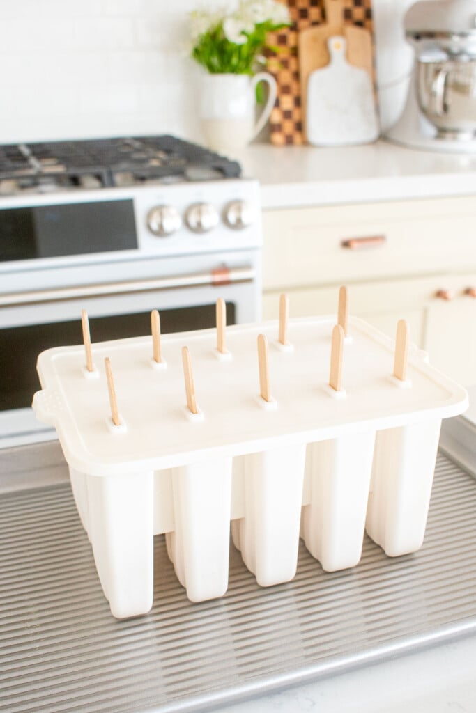 orange creamsicle popsicle mixture in molds with sticks on a baking sheet on a marble counter.