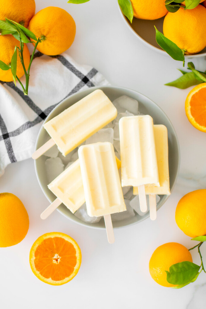 orange creamsicle popsicles in a gray bowl on a white marble counter with fresh oranges.