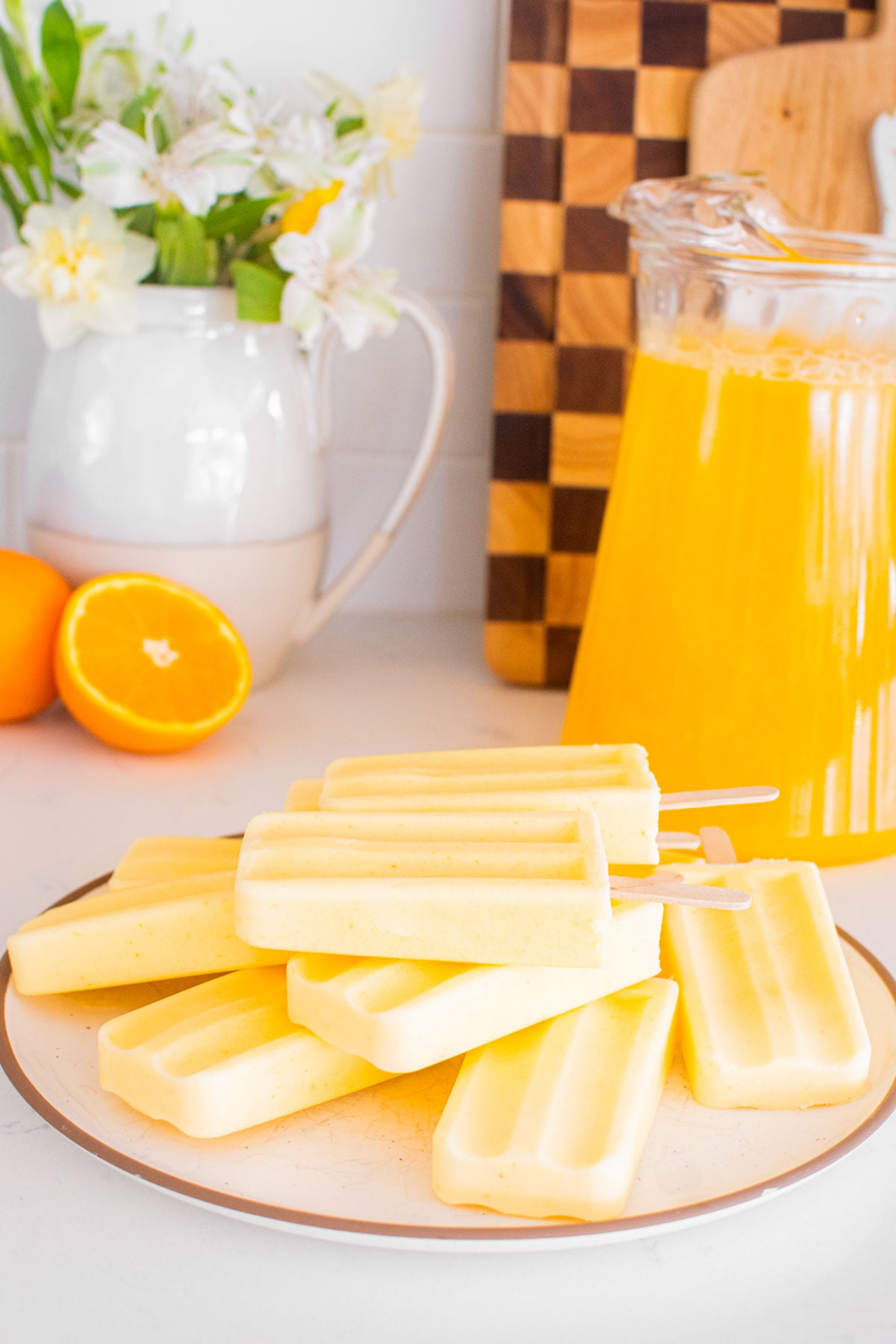 orange creamsicle popsicles on a plate on a white marble kitchen counter with fresh oranges and orangeade in a pitcher.