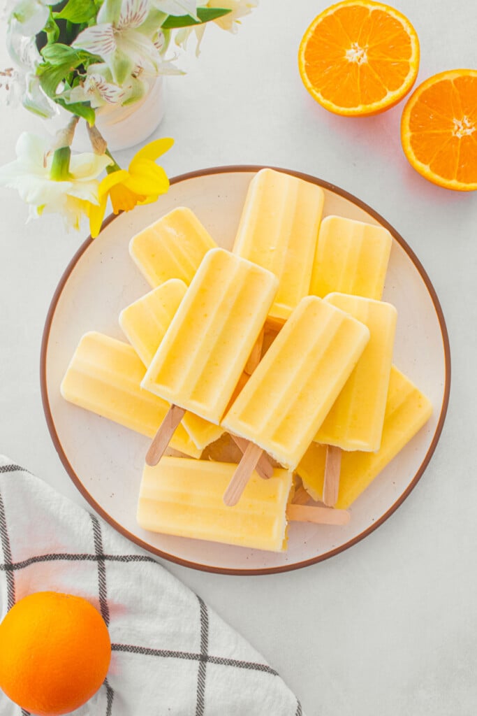 orange creamsicle popsicles on a plate on a table with fresh oranges and flowers.