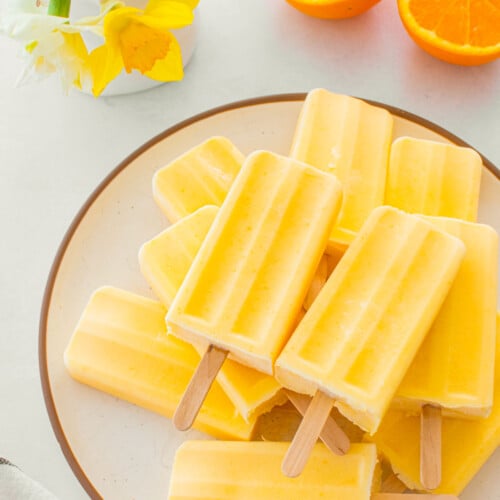 orange creamsicle popsicles on a plate on a table with fresh oranges and flowers.