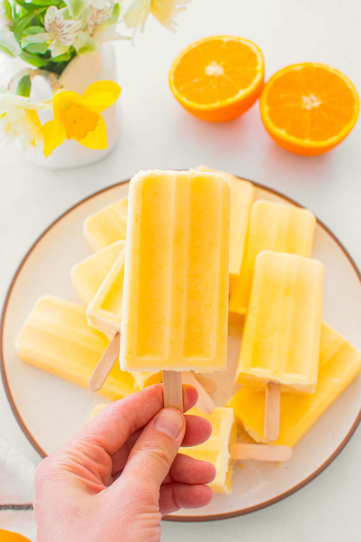 woman holding an orange creamsicle popsicle above a plate of popsicles with fresh oranges and flowers.