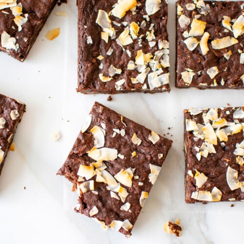 vegan toasted coconut brownies on a white marble counter, one brownie with a bite taken out.