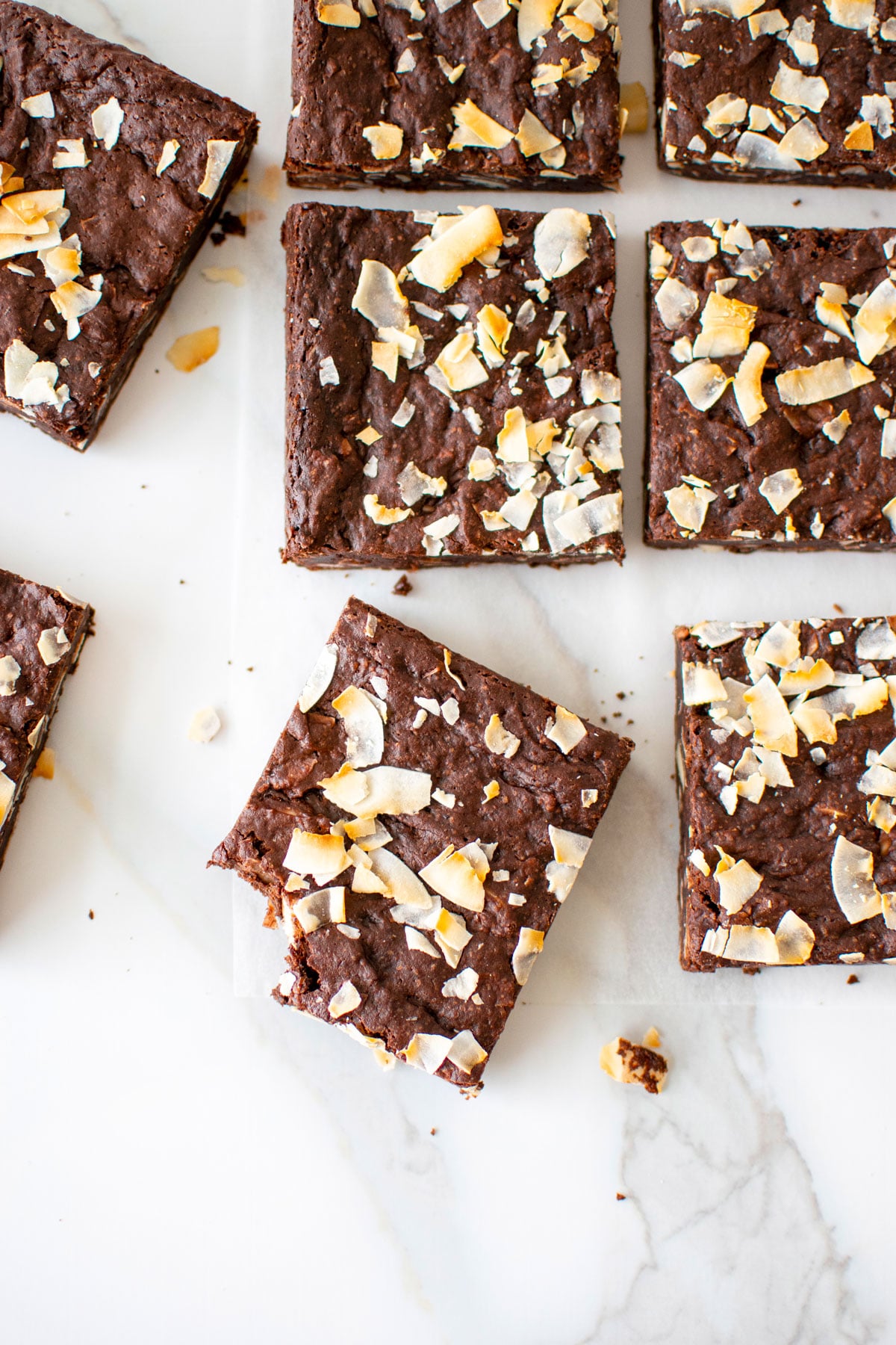 vegan toasted coconut brownies on a white marble counter, one brownie with a bite taken out.