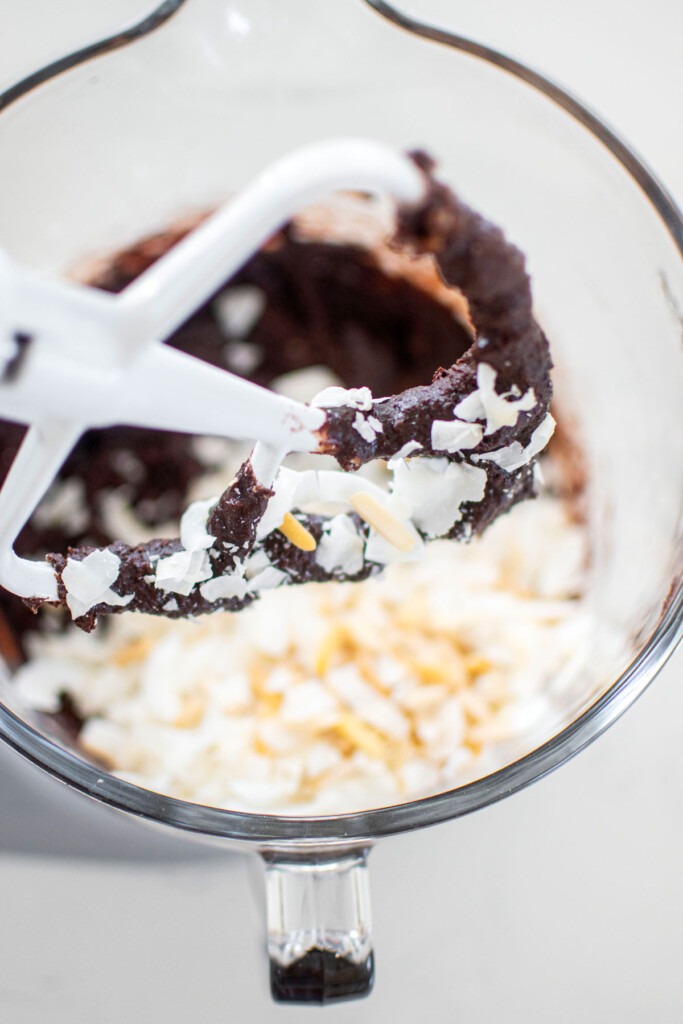 vegan toasted coconut brownie batter with coconut in it in a glass mixing bowl on a white marble counter.