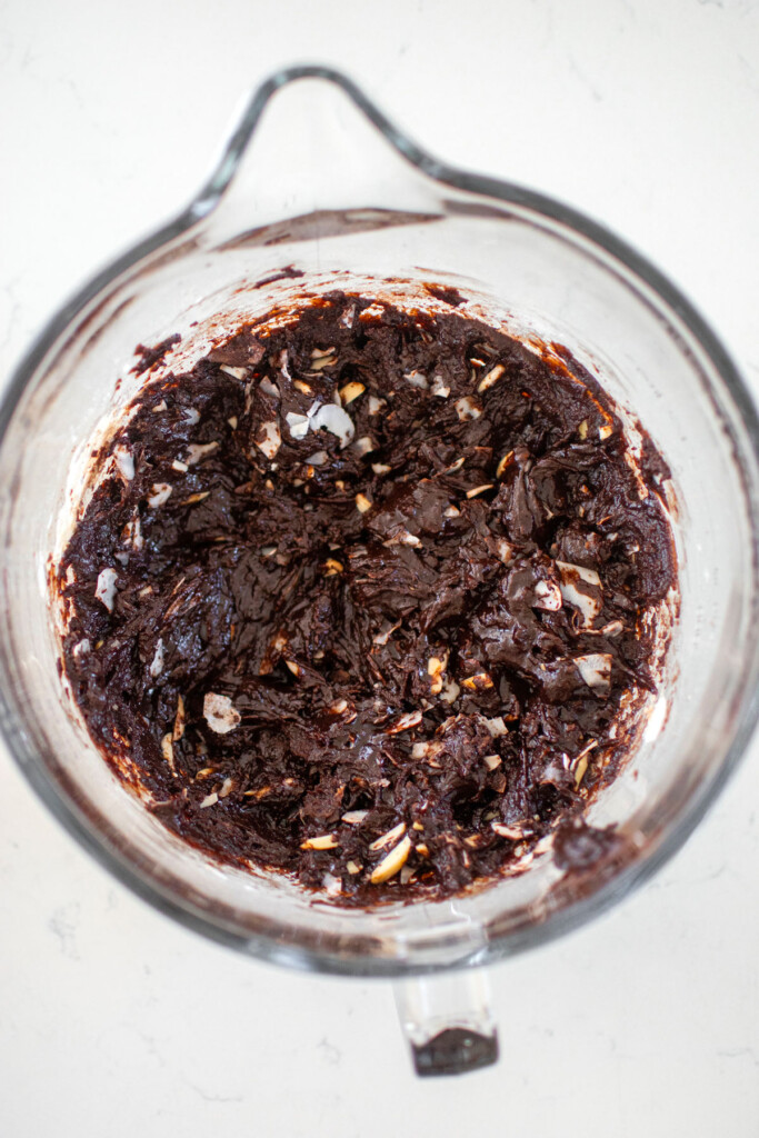 vegan toasted coconut brownie batter in a glass mixing bowl on a white marble counter.
