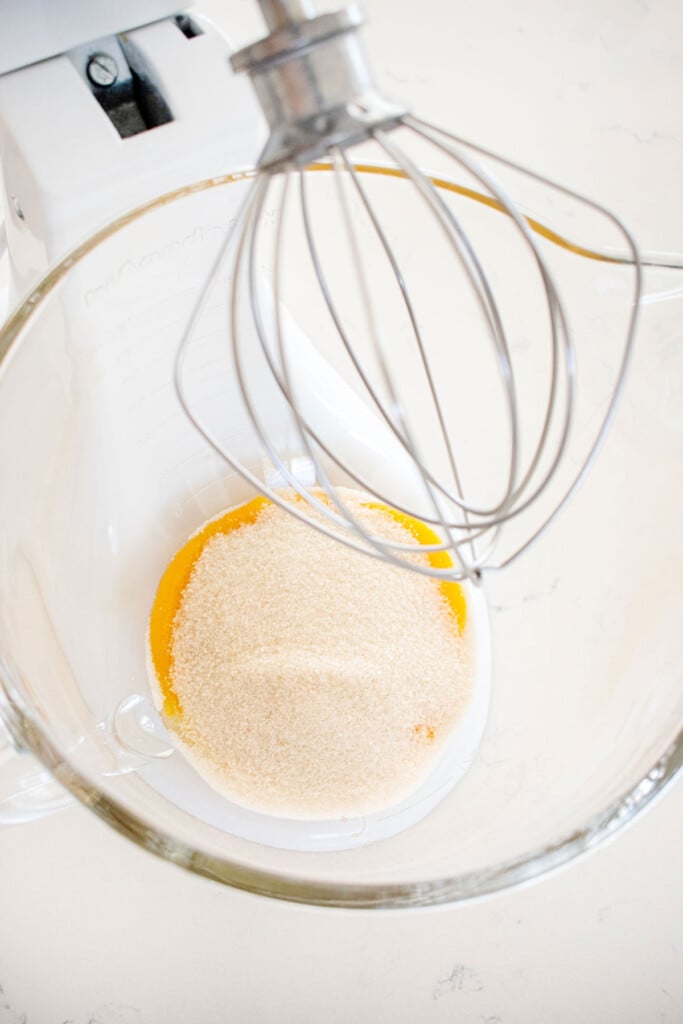 egg yolks and sugar in a glass mixing bowl with a whisk on a white marble counter.