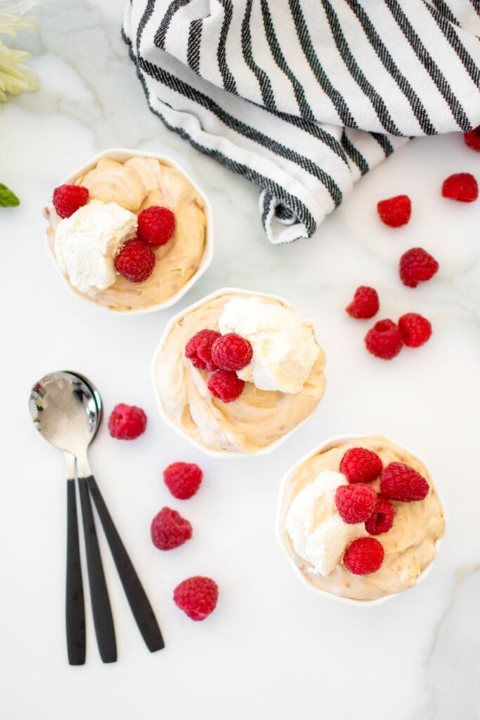 fresh raspberry custard in small white bowls with homemade whipped cream and fresh raspberries on a marble counter.