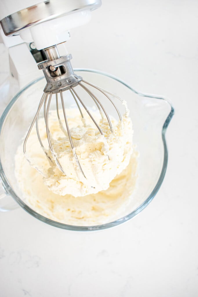 homemade whipped cream in a glass mixing bowl on a white marble counter.