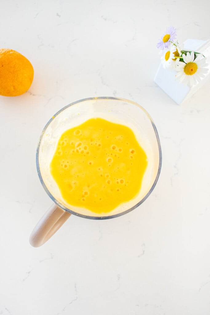 blender with blended orange julius on a marble counter with an orange and fresh flowers.