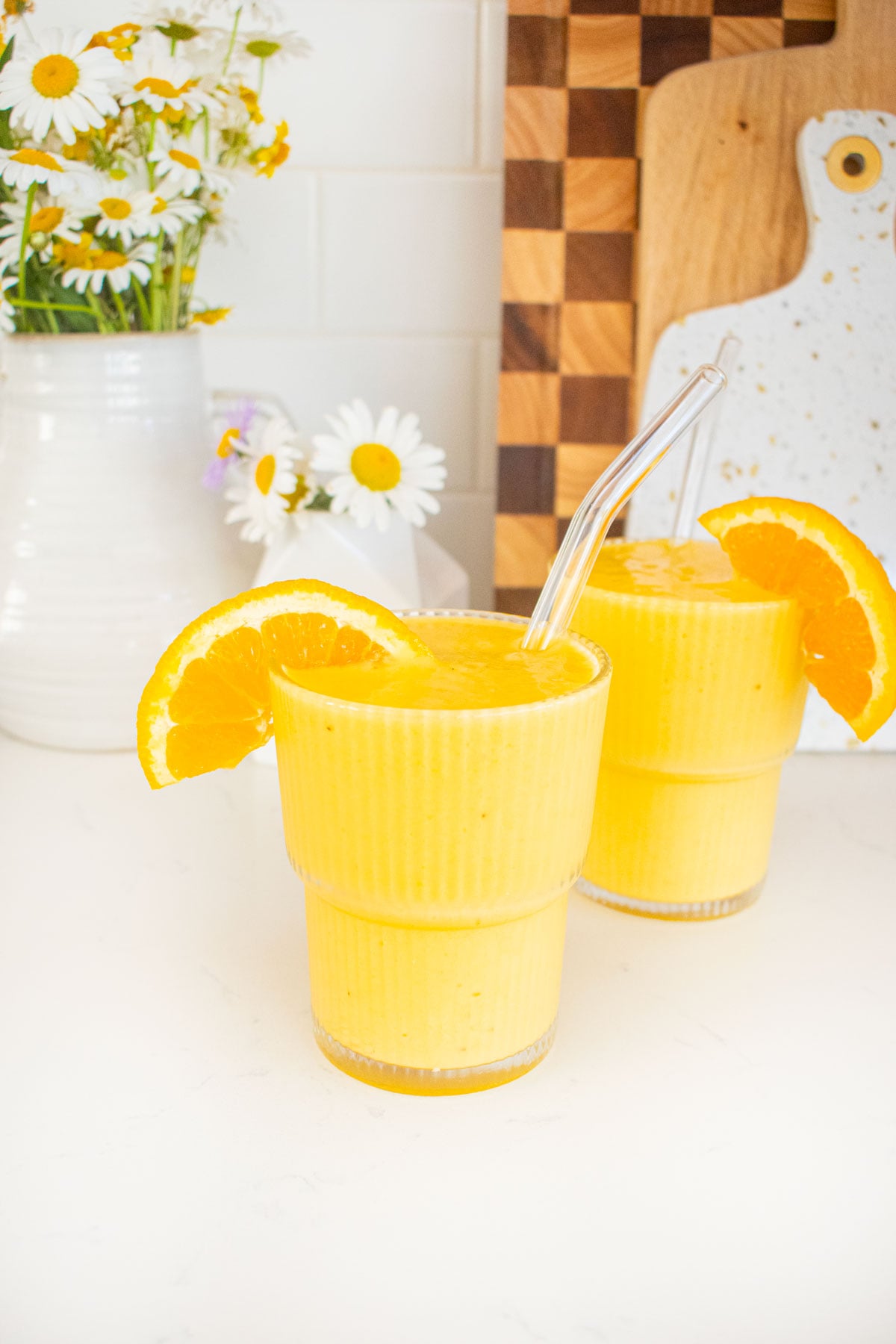two glasses of orange julius with fresh orange slices and straws on a marble counter with fresh flowers.