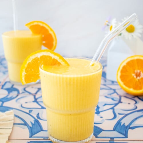 two glasses of orange julius with an orange slice on the side and straws on a blue tile table with fresh flowers.