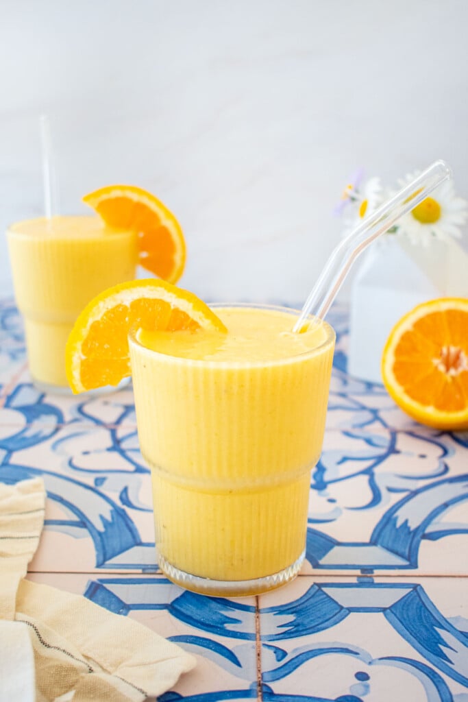 two glasses of orange julius with an orange slice on the side and straws on a blue tile table with fresh flowers.