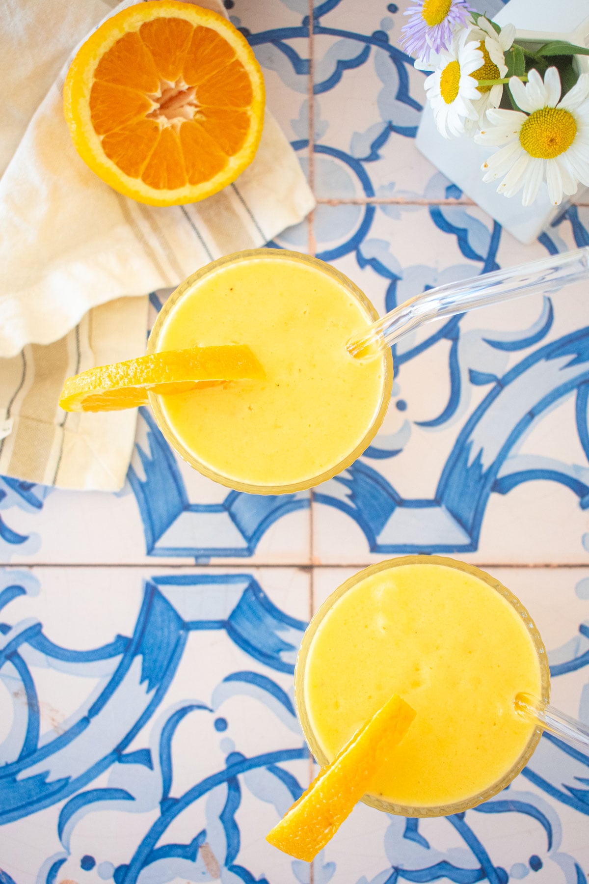 overhead view of two glasses of orange julius with an orange slice on the side and straws on a blue tile table with fresh flowers.