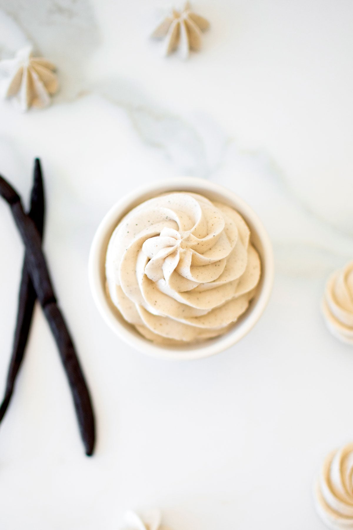 vanilla bean buttercream frosting swirled in a small bowl on a white marble counter with vanilla bean pods.