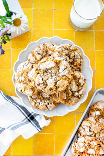 plate of chocolate chip crinkle cookies on a yellow tile counter with a glass of milk and fresh flowers.