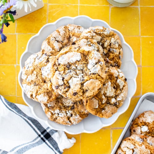 plate of chocolate chip crinkle cookies on a yellow tile counter with a glass of milk and fresh flowers.
