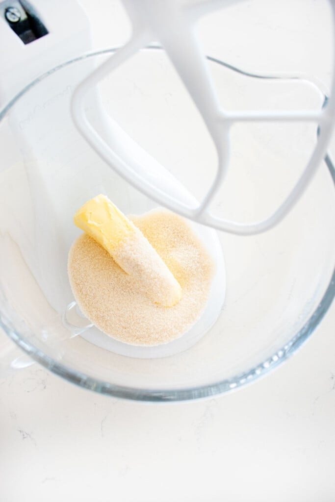butter and cane sugar in a glass stand mixing bowl on a white marble counter.