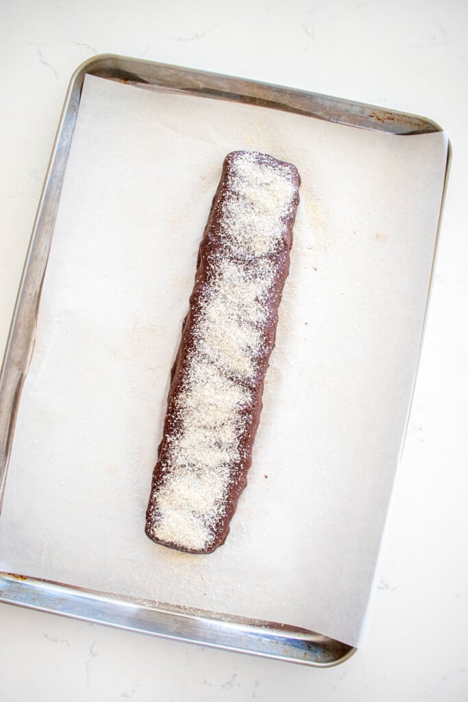 chocolate malt biscotti dough log on a baking sheet sprinkled with cane sugar on a marble counter.