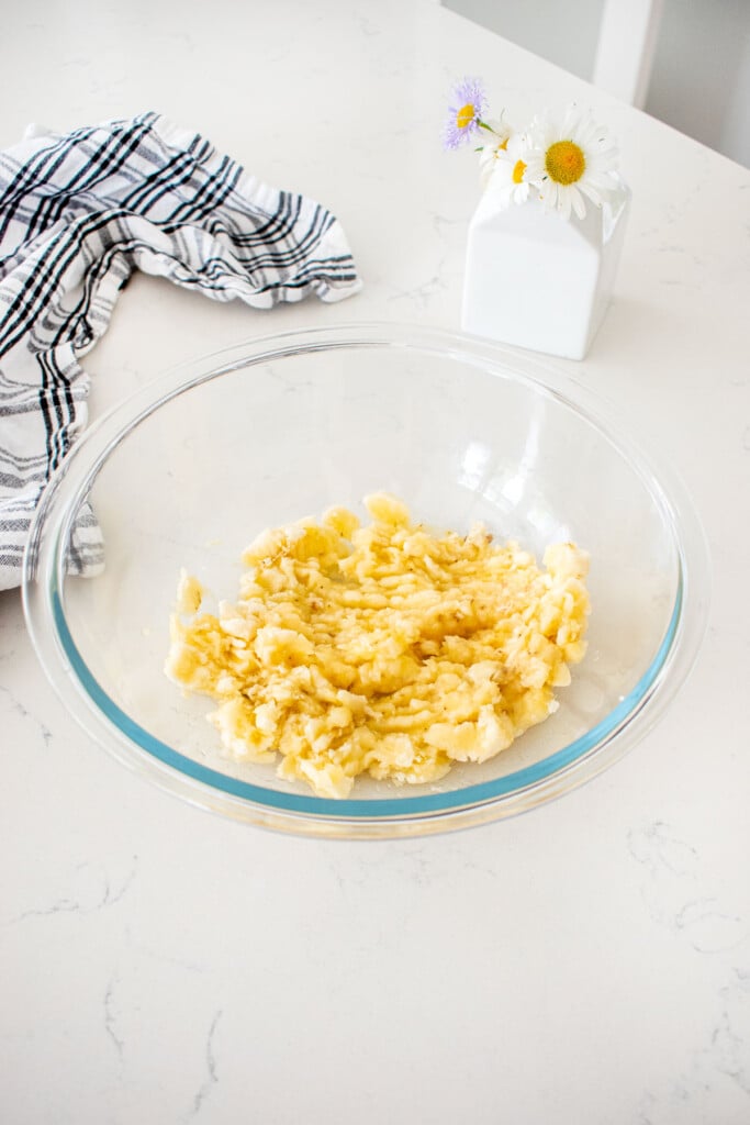 glass mixing bowl with mashed bananas on a marble counter with a vase of fresh flowers.