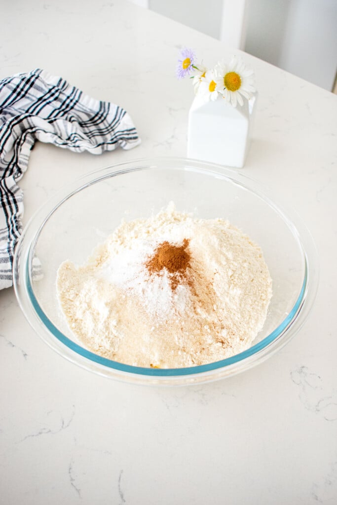 glass mixing bowl with ingredients for eggless banana waffles on a marble counter with a vase of fresh flowers.