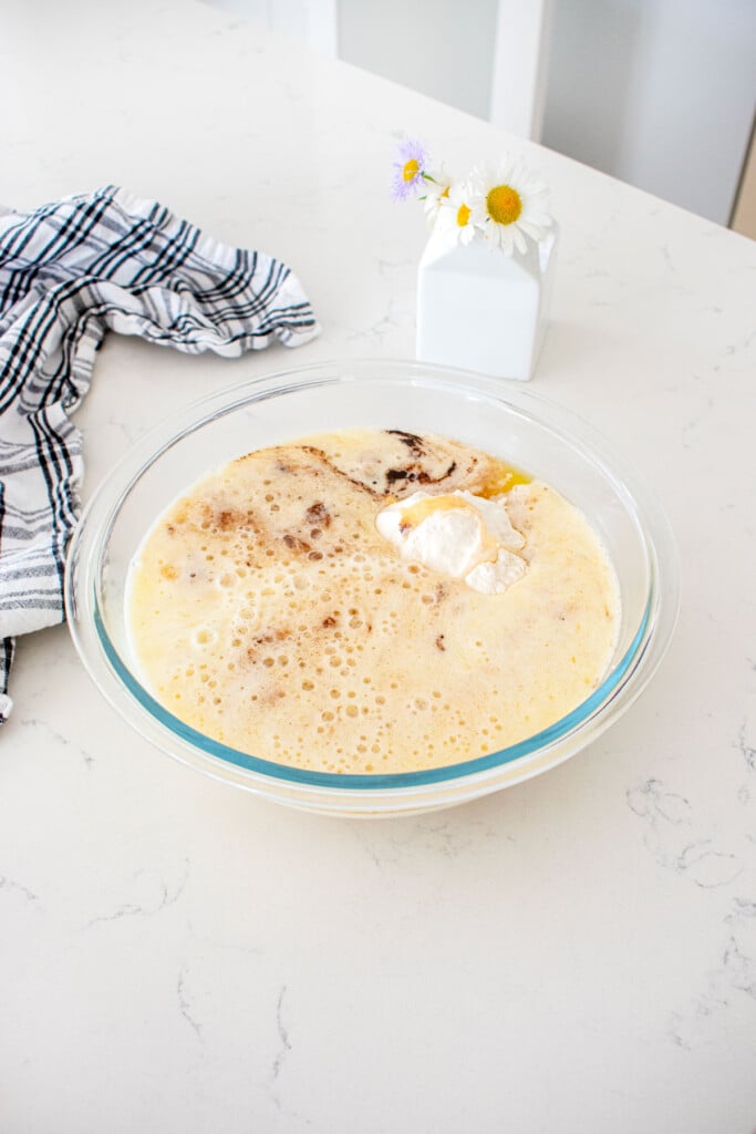 glass mixing bowl with ingredients for eggless banana waffles on a marble counter with a vase of fresh flowers.
