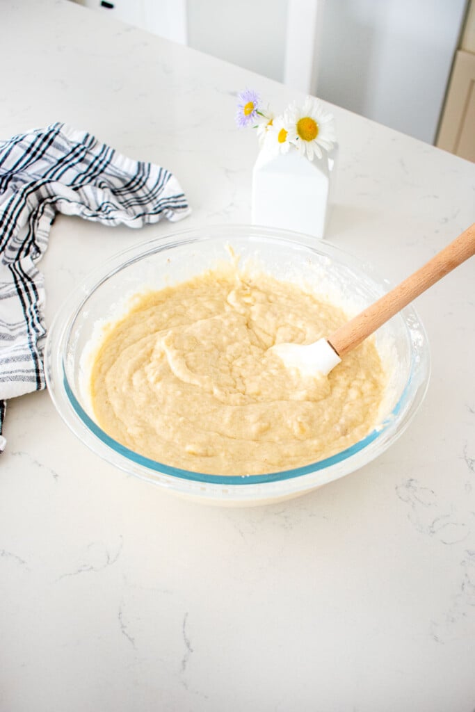 eggless banana waffle batter in a glass mixing bowl with a spatula on a marble counter with a vase of flowers.