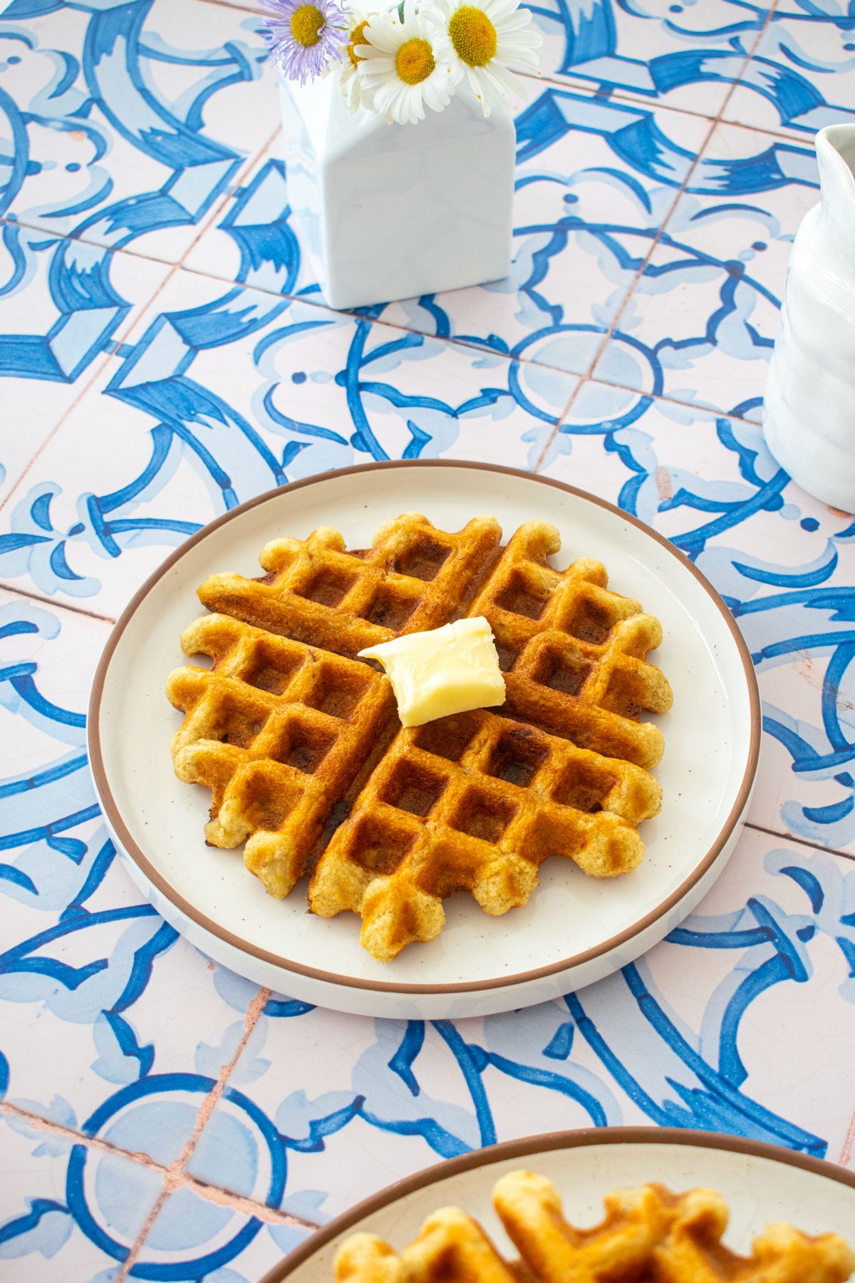 golden brown eggless banana waffle on a plate with a pad of butter on a tile table with a vase of flowers.