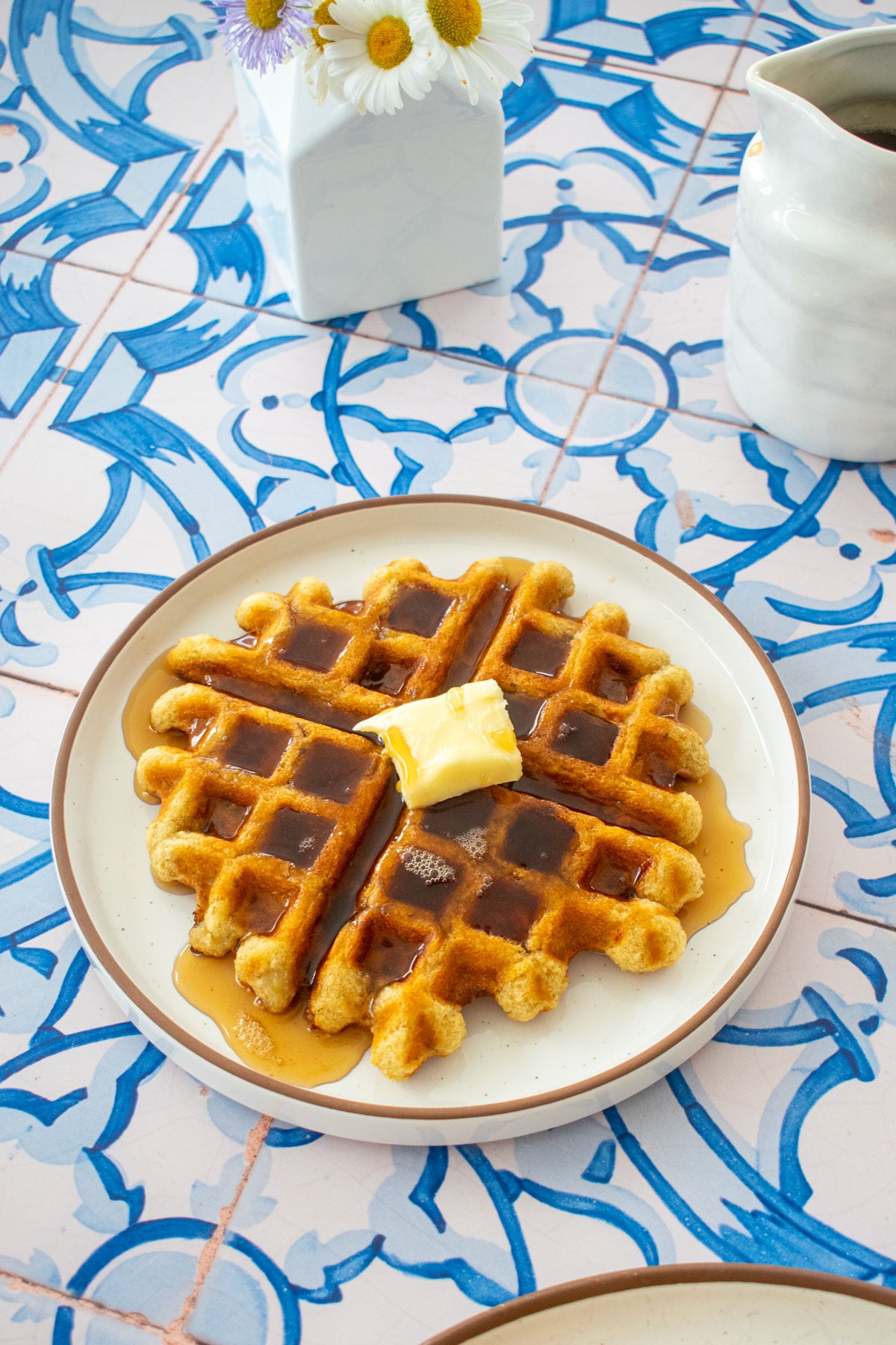 golden brown eggless banana waffle on a plate with a pad of butter and maple syrup on a tile table with fresh flowers.