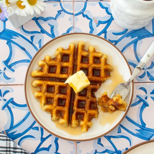 golden brown eggless banana waffle on a plate with a pad of butter and maple syrup on a tile table with fresh flowers.