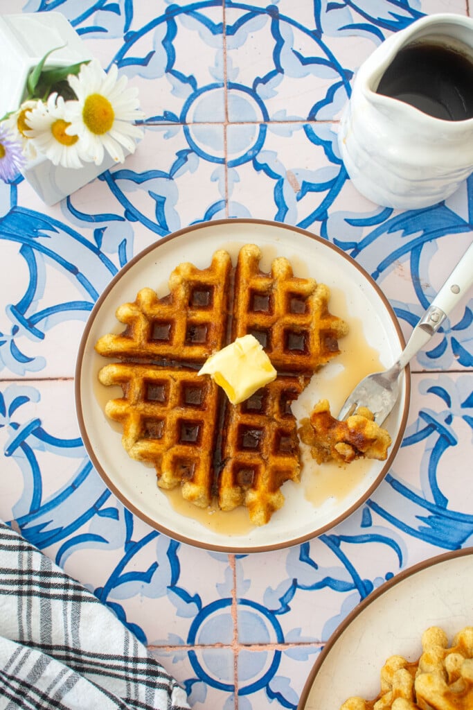 golden brown eggless banana waffle on a plate with a pad of butter and maple syrup on a tile table with fresh flowers.