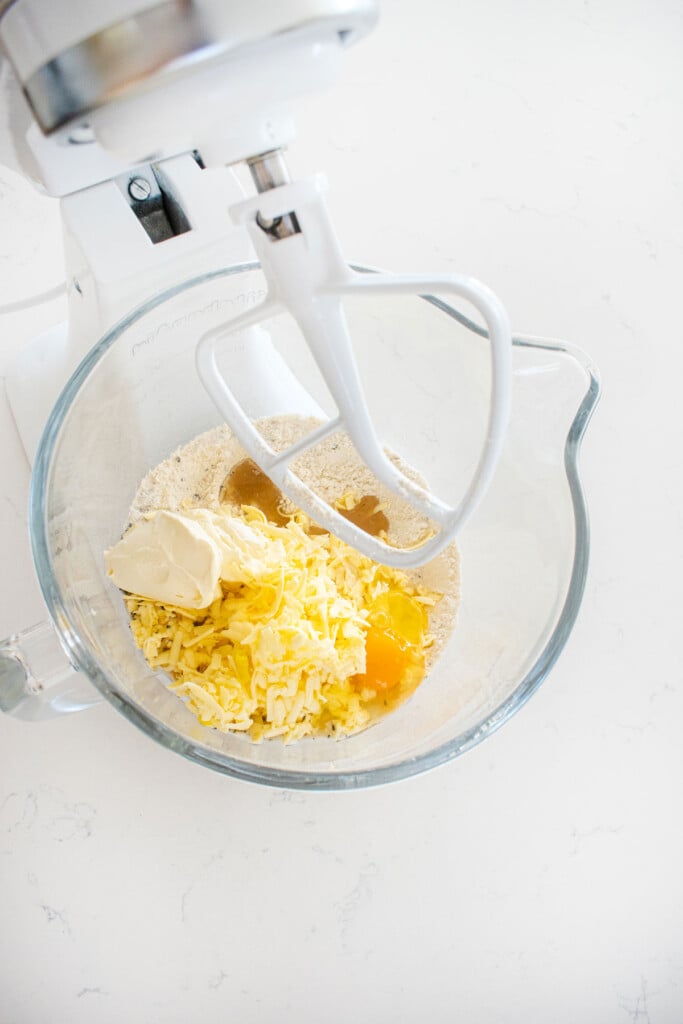 flour, grated butter, egg and cream cheese in a glass mixing bowl on a white countertop.