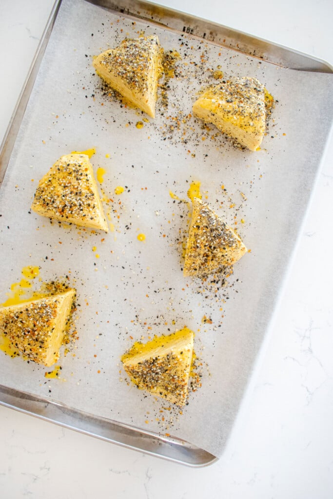 everything bagel scones on a baking sheet brushed with egg wash and everything seasoning.